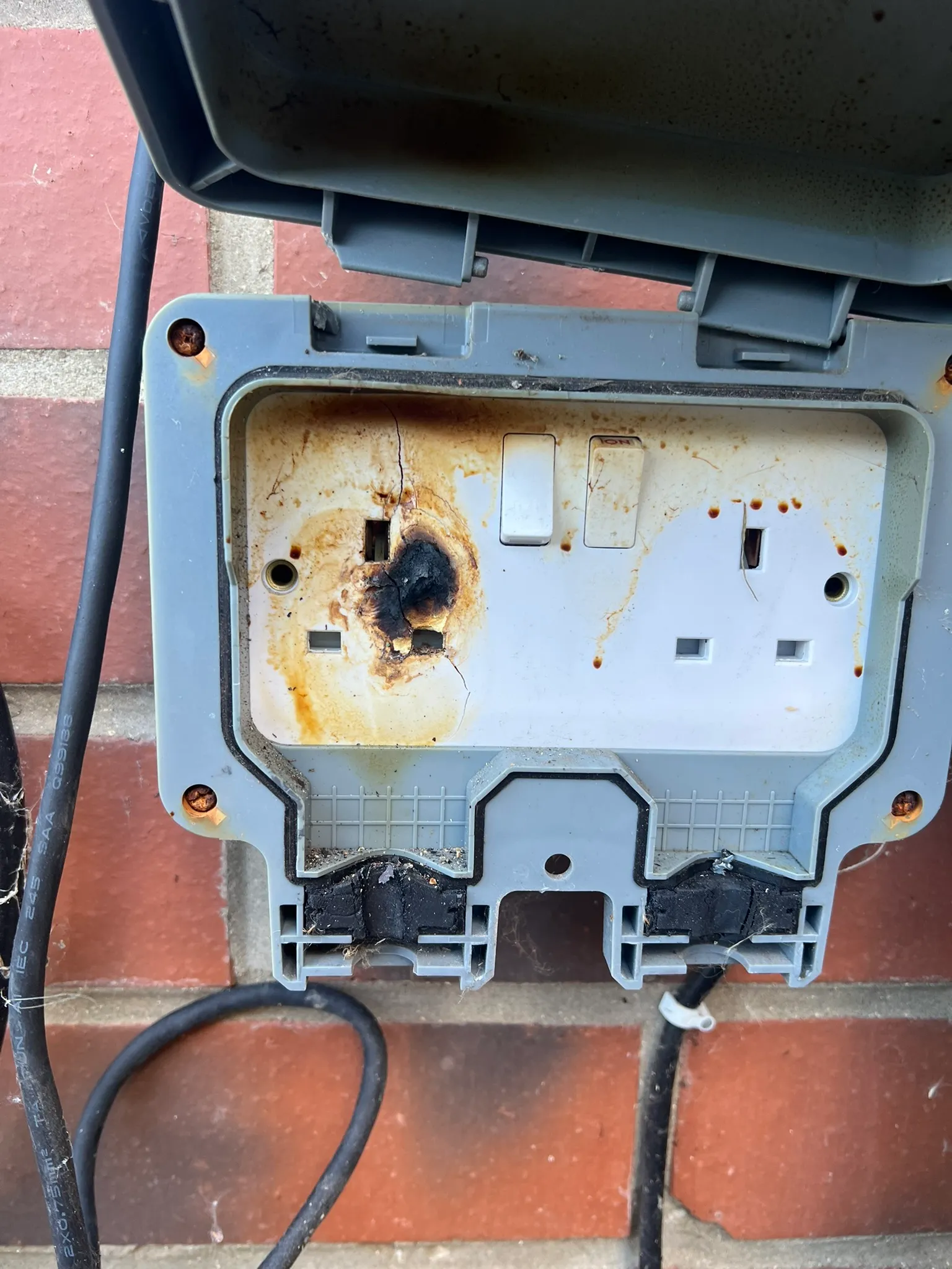 Outdoor double socket with a charred, cracked faceplate around one set of pin holes and brown staining