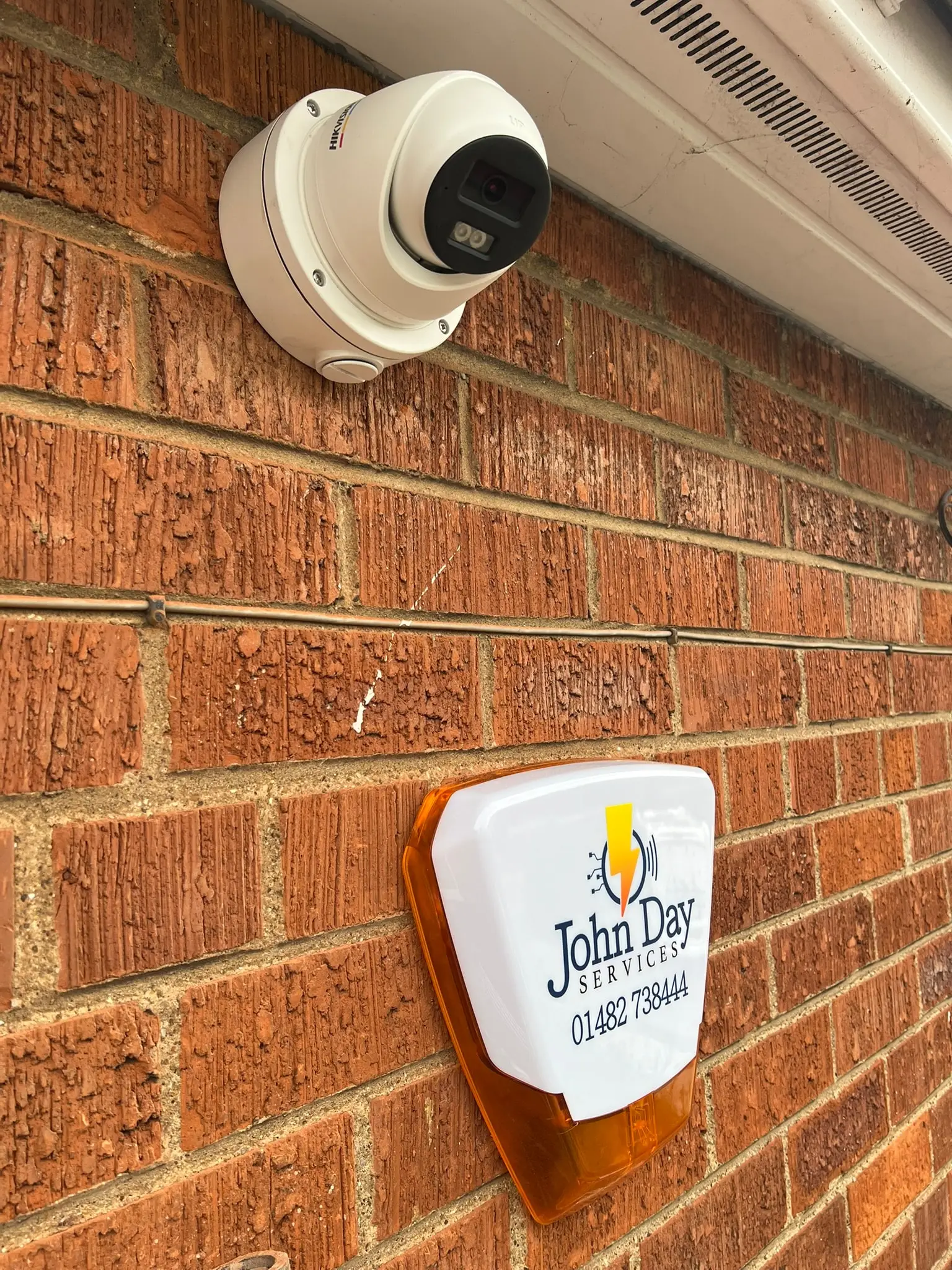 A white Hikvision dome camera under the eaves and a John Day Services alarm bell box on a brick wall, both installed by us