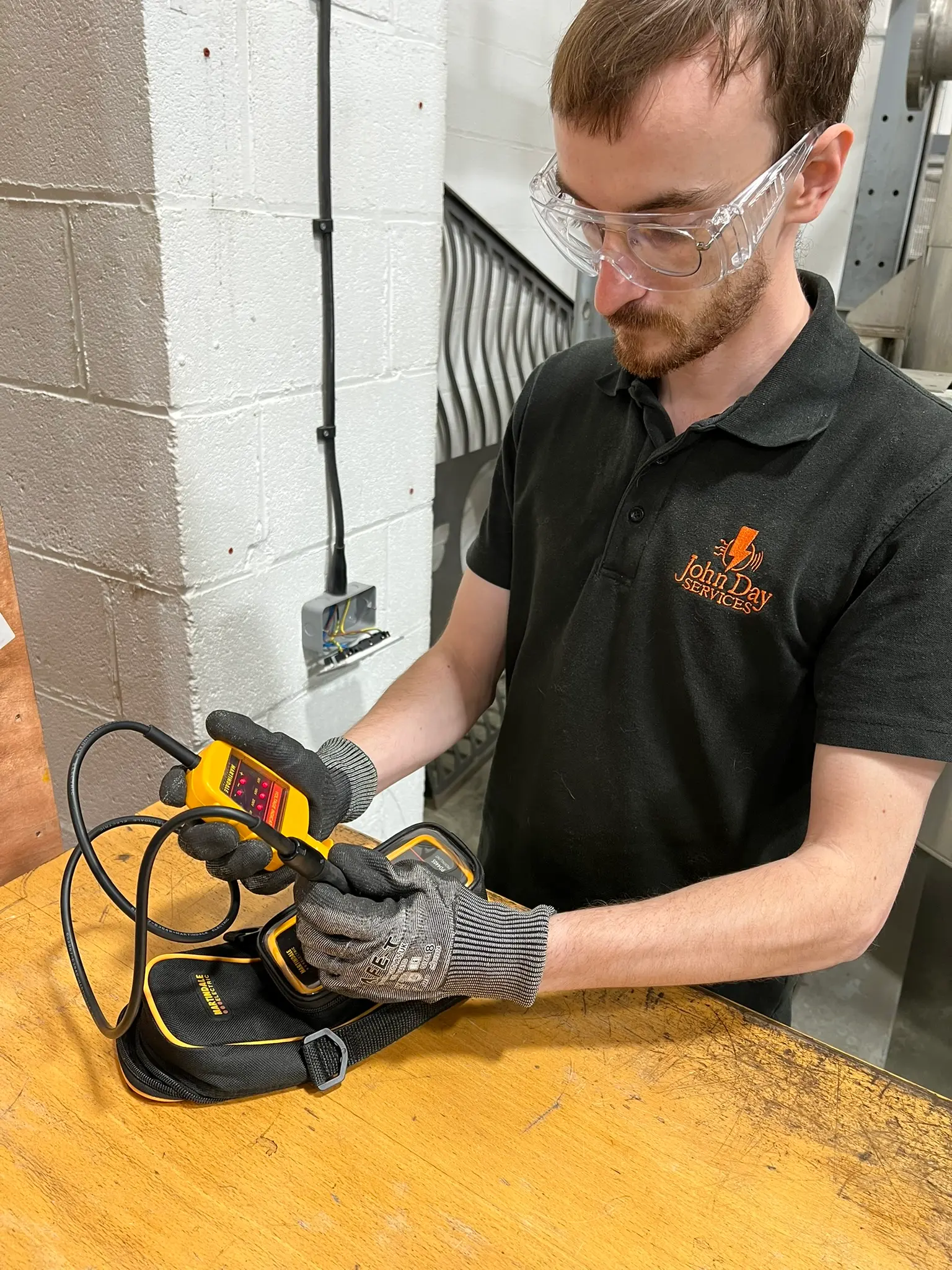 Joe in safety glasses and a John Day Services polo shirt, checking a voltage tester