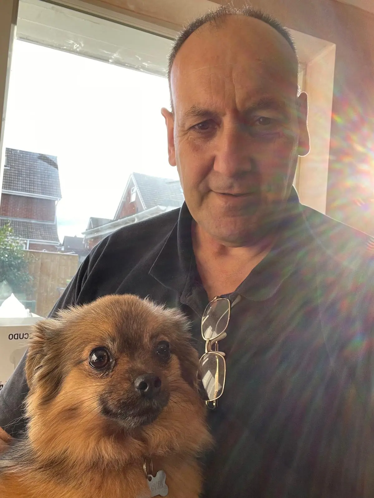 John in a dark polo shirt, holding a small fluffy brown dog by a sunny window