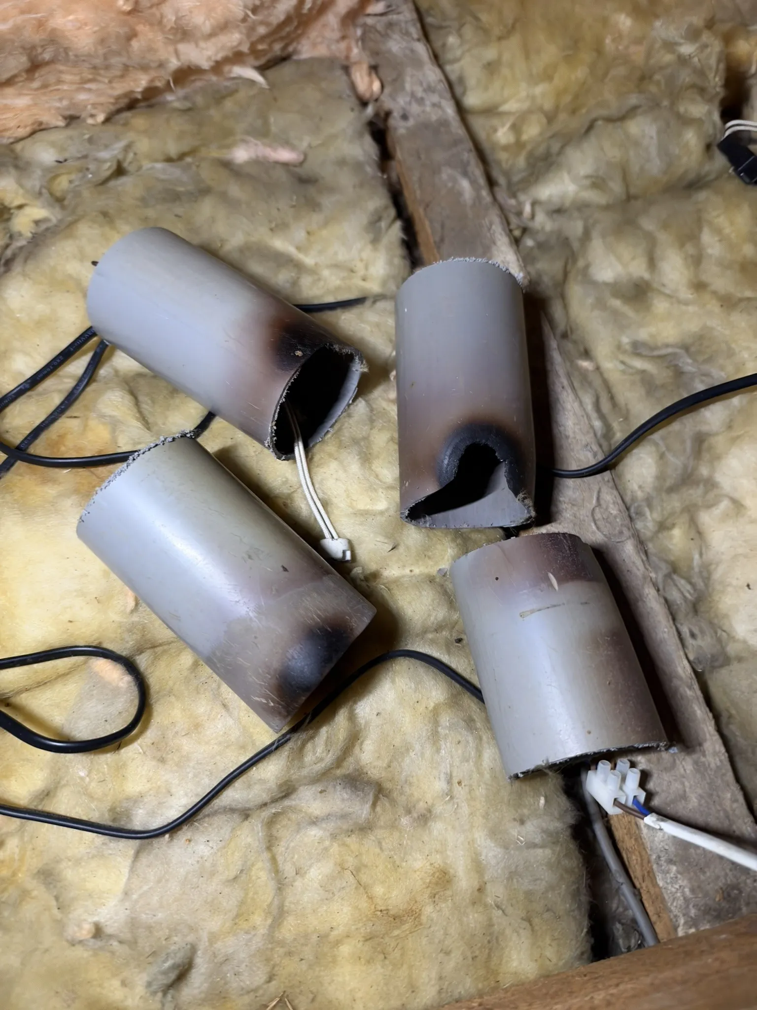 Four sections of grey plastic pipe on loft insulation, scorched black at one end, one with a hole burnt through, with cables running between them