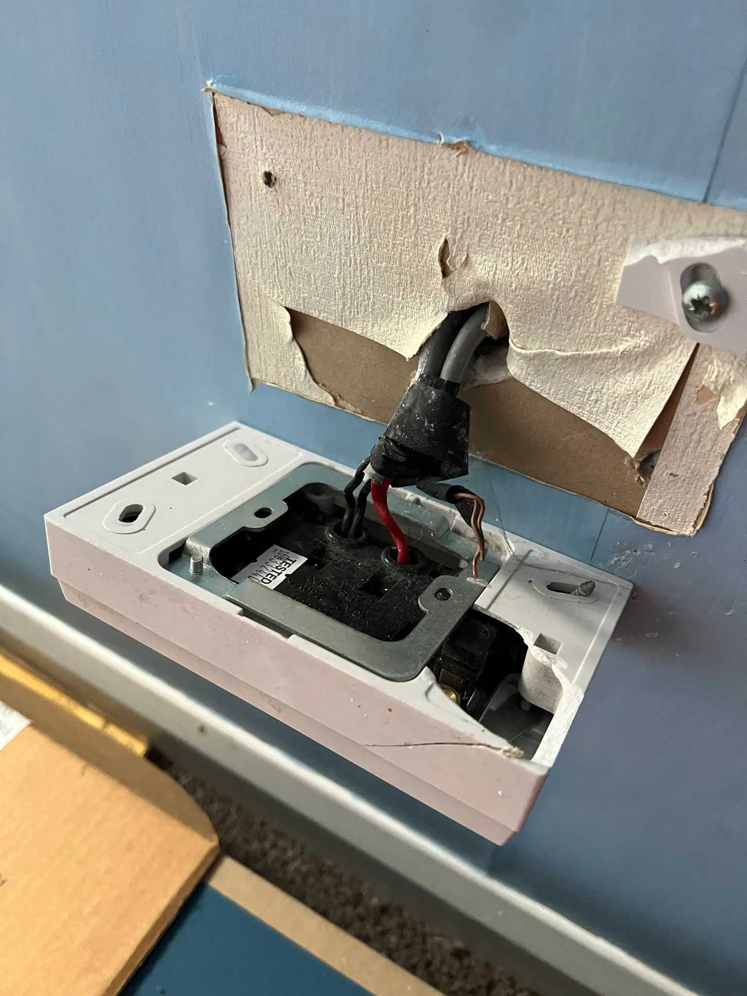 Cracked socket box hanging from a torn opening in plasterboard, with taped cables